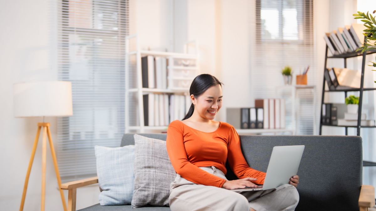 Asian woman using her laptop while relaxing on a sofa in a bright living room, enjoying Usenet.