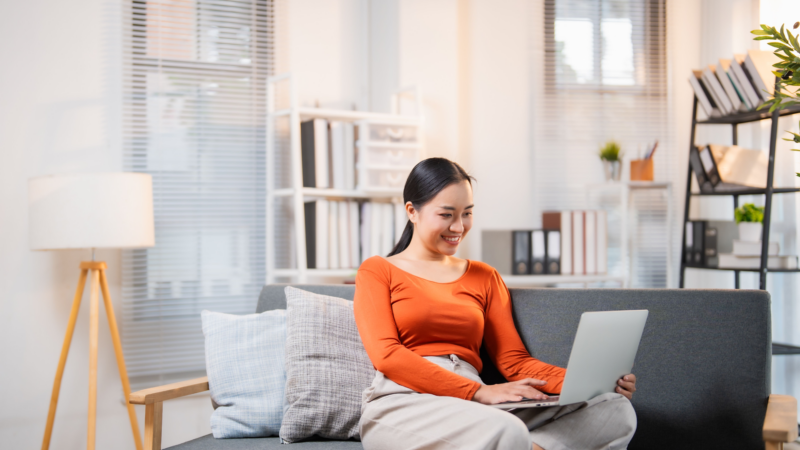 Asian woman using her laptop while relaxing on a sofa in a bright living room, enjoying Usenet.